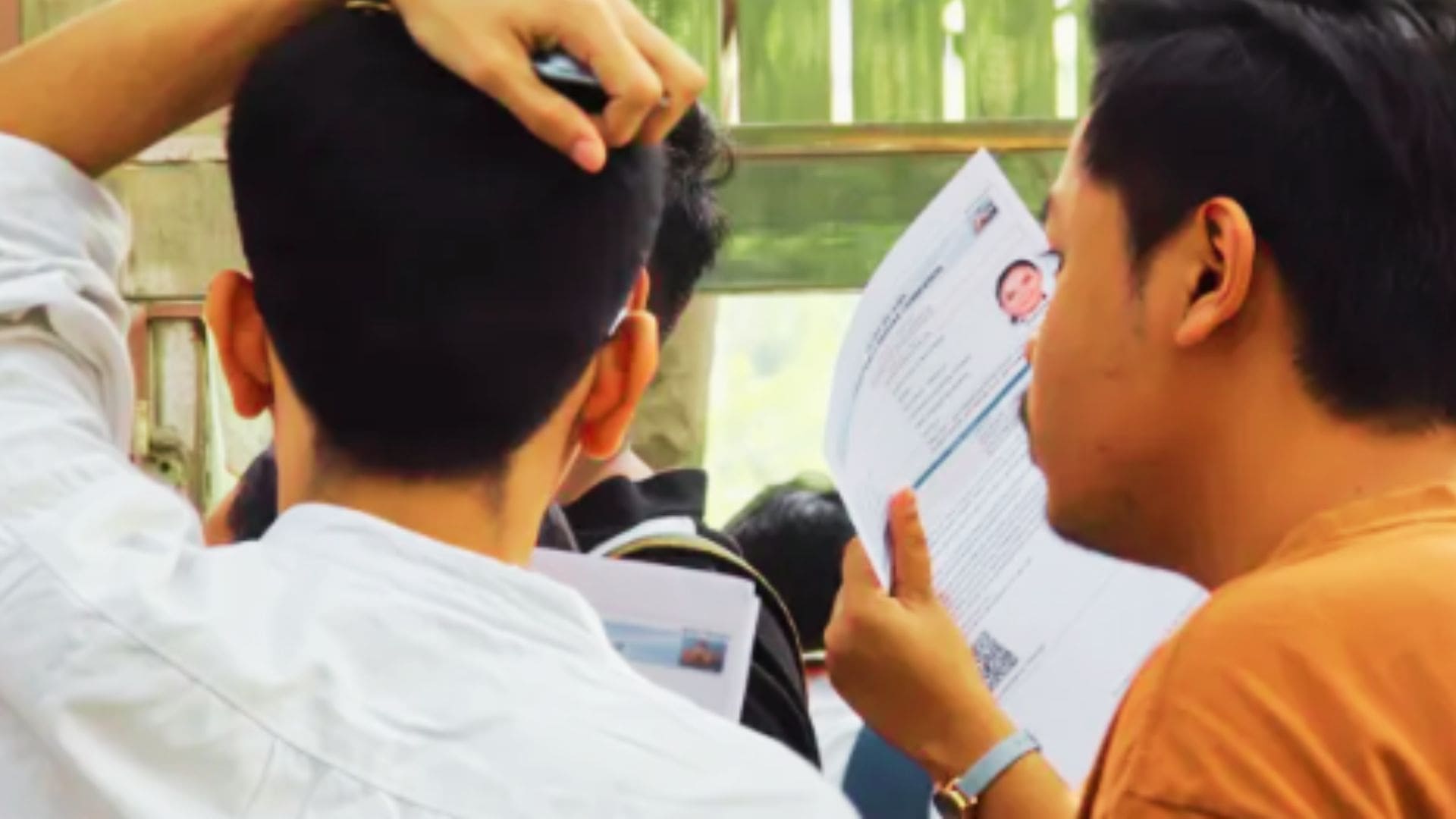 A group of students standing in a queue, with one student holding a document containing a passport-size photo while another scratches their head, conveying anticipation and nervousness during a registration, admission, or document verification process.