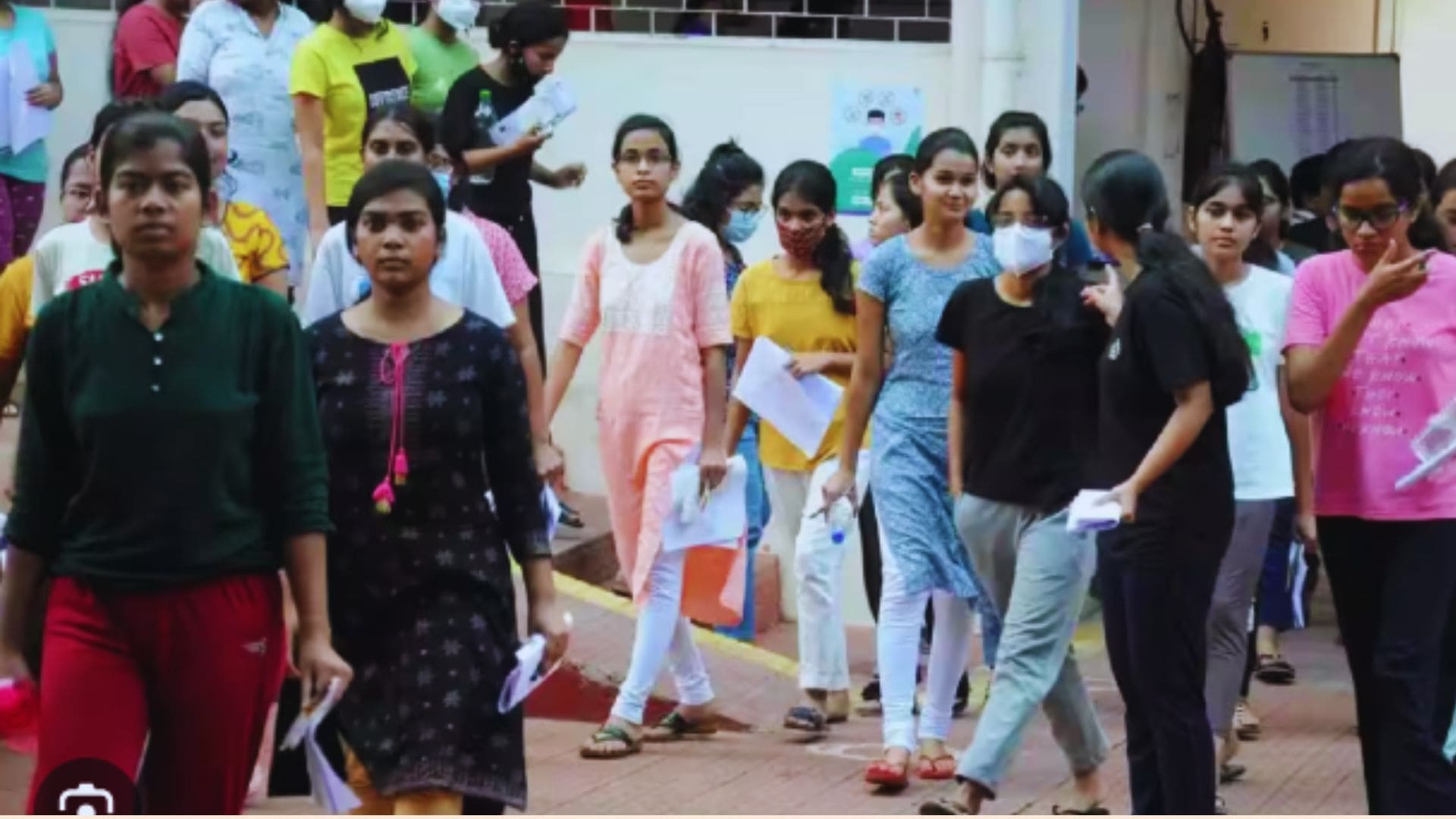 A group of students walk out of an examination center holding question papers and documents after completing an exam. Some students are talking with friends, while others appear focused as they leave the campus. Several students are wearing face masks, and the scene captures the atmosphere immediately after an exam.