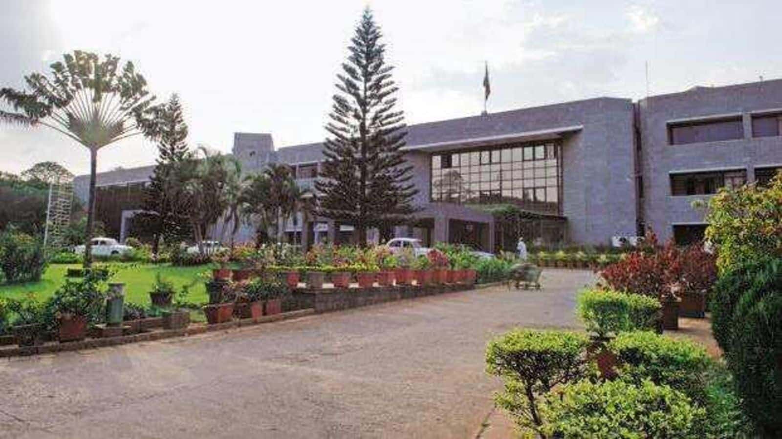 Exterior view of the ISRO headquarters building featuring a modern grey facade with large glass windows, a landscaped garden with lush greenery, potted plants, tall trees, and a wide driveway leading to the main entrance under a bright daytime sky.